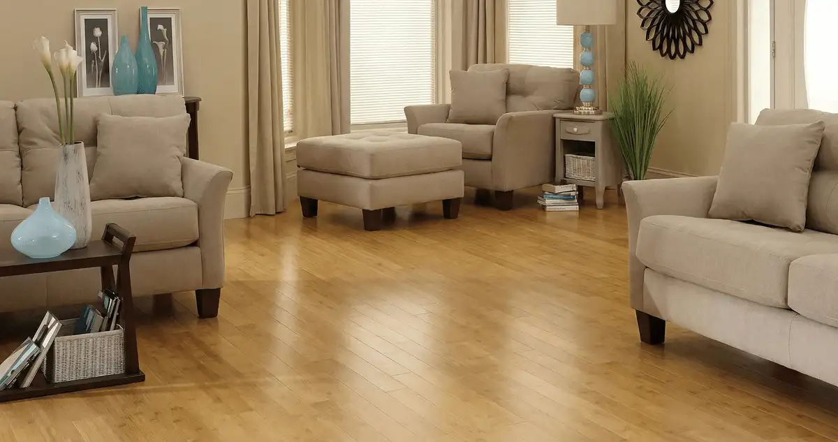 A living room with professionally installed bamboo flooring, beige sofas, a matching ottoman, a side table with a blue lamp, vases, framed pictures, and large windows with blinds and curtains in Fort Lauderdale, FL